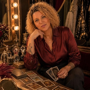 May include: A person with curly blonde hair wearing a long-sleeved, button-down, dark red satin shirt. The person is seated at a wooden table with several tarot cards spread out, holding one card in their hand. A vanity mirror with lights is visible.