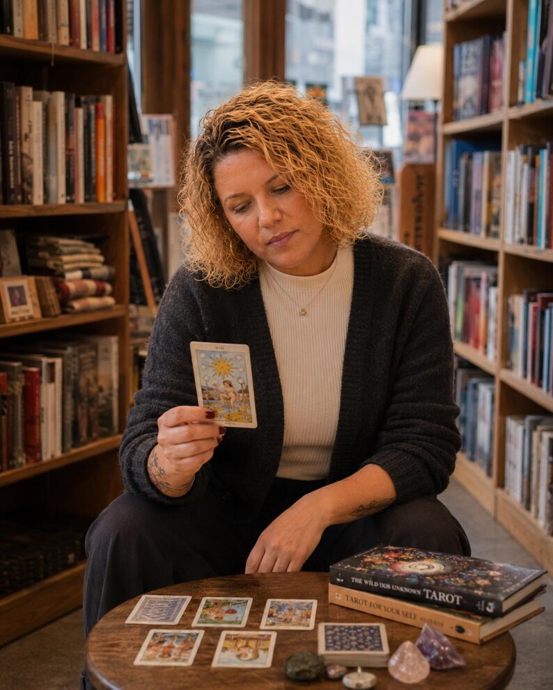 May include: A person holding a tarot card featuring a sun illustration, wearing a dark gray cardigan over a white shirt. On the wooden table are several tarot cards, a deck of cards, and two books titled The Wild Unknown Tarot and Tarot For Your Self.