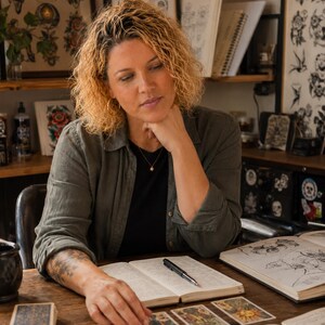 Peut inclure: Une personne aux cheveux blonds boucl&eacute;s portant une chemise boutonn&eacute;e vert olive sur un haut noir, assise &agrave; une table en bois avec un carnet ouvert, un stylo noir et un jeu de cartes de tarot dispos&eacute; en rang&eacute;e, avec des dessins de tatouage en arri&egrave;re-plan.