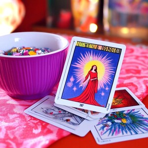 Pu&ograve; includere: Un'apertura di carte dei tarocchi su una superficie con motivo rosa e rosso. La carta superiore &egrave; la Sacerdotessa, che mostra una donna con una veste rossa in piedi davanti a un raggio di sole. La carta &egrave; circondata da altre carte, tra cui una con un design blu e verde.