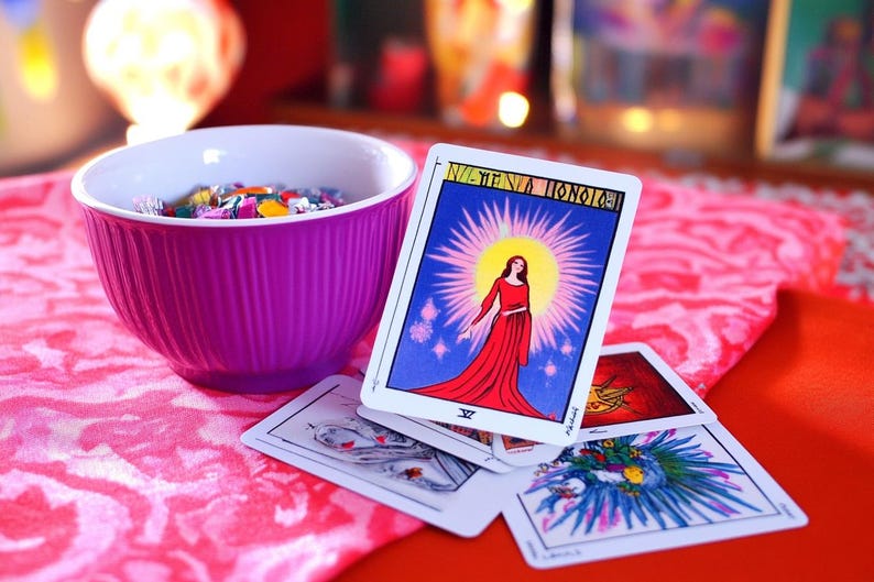 Pu&ograve; includere: Un'apertura di carte dei tarocchi su una superficie con motivo rosa e rosso. La carta superiore &egrave; la Sacerdotessa, che mostra una donna con una veste rossa in piedi davanti a un raggio di sole. La carta &egrave; circondata da altre carte, tra cui una con un design blu e verde.
