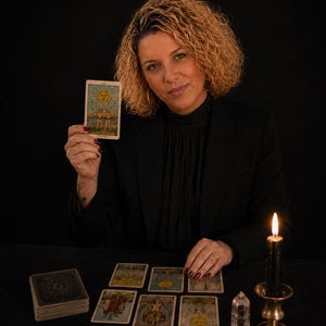 May include: A tarot card titled The Sun featuring a yellow sun and two figures, held up by a person in a black blazer. On the table are several tarot cards, a deck of cards, a clear crystal point, and a black candle in a brass holder.