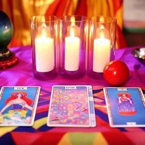Pu&ograve; includere: Tre candele accese in portacandele di vetro, una mela rossa, una sfera di cristallo su un supporto e tre carte dei tarocchi su una tovaglia da tavolo colorata. Le carte sono etichettate con "IV", "XVII" e "XI".