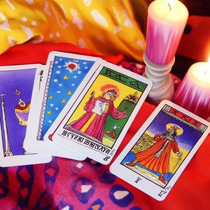 May include: Four tarot cards, including the Eight of Wands, the Ace of Cups, the Tower, and the King of Wands, are spread out on a red and yellow patterned fabric. Two lit candles are in the background.