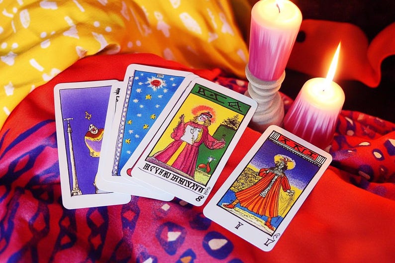 May include: Four tarot cards, including the Eight of Wands, the Ace of Cups, the Tower, and the King of Wands, are spread out on a red and yellow patterned fabric. Two lit candles are in the background.