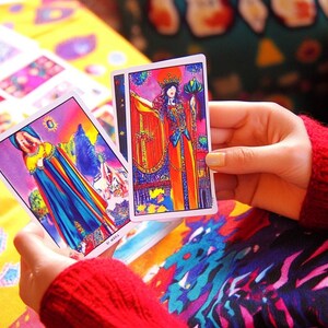 Pu&ograve; includere: Due carte dei tarocchi, una con una donna in una veste blu e arcobaleno e l'altra con una donna in una veste rossa e arancione, sono tenute nelle mani di una persona.