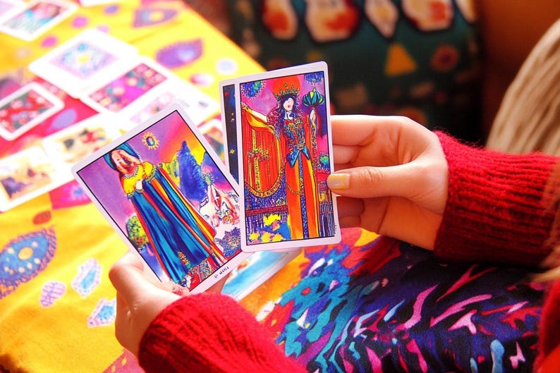 Pu&ograve; includere: Due carte dei tarocchi, una con una donna in una veste blu e arcobaleno e l'altra con una donna in una veste rossa e arancione, sono tenute nelle mani di una persona.
