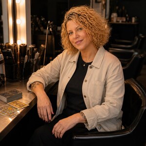 May include: A person with curly blonde hair wearing a beige button-down jacket over a black shirt, seated in a black chair. On the table are several tarot cards with intricate gold and blue designs, alongside a stack of cards and hair styling tools.