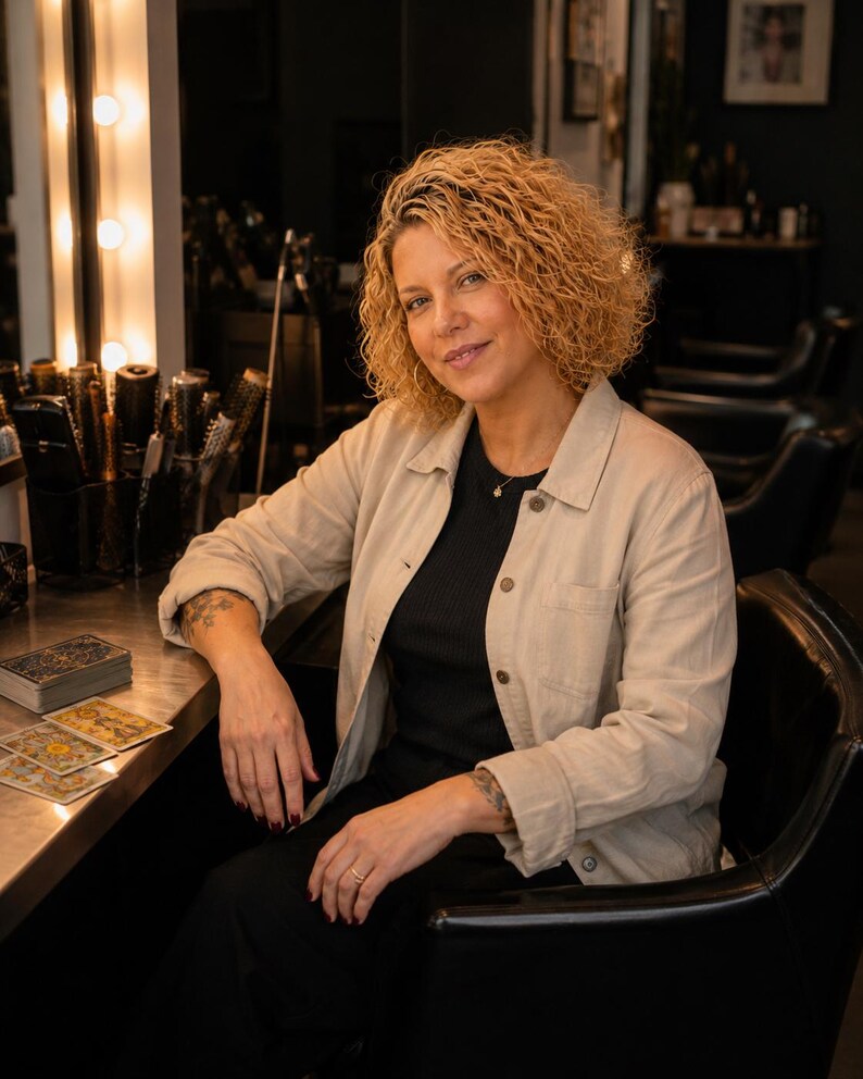 May include: A person with curly blonde hair wearing a beige button-down jacket over a black shirt, seated in a black chair. On the table are several tarot cards with intricate gold and blue designs, alongside a stack of cards and hair styling tools.