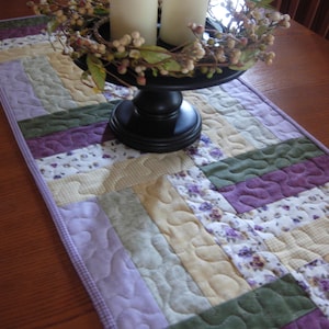 May include: A quilted table runner with a patchwork design in shades of lavender, green, and cream. A black pedestal holds two white candles and a decorative floral wreath. The runner is approximately 30 cm wide.