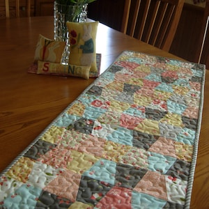 May include: A quilted table runner with a geometric pattern in shades of grey, blue, pink, and yellow. The runner has a white and grey striped border. The runner is on a wooden table with a vase of flowers and decorative cushions.