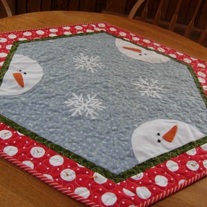 May include: A blue and white quilted table runner featuring a snowflake and snowman design. The runner has a red and white polka dot border with a green and white trim.