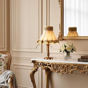 May include: A decorative gold mirror above a gold console table with a marble top. A floral armchair and a lamp with a fringed shade are also in the room. The room has a classic, elegant style.