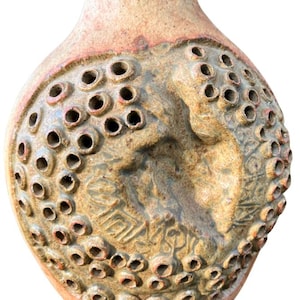 Puede incluir: Lámpara colgante de cerámica hecha a mano. La lámpara tiene una combinación de colores beige y marrón rojizo, con una superficie texturizada y numerosos agujeros pequeños. Un diseño en relieve es visible en la parte delantera.