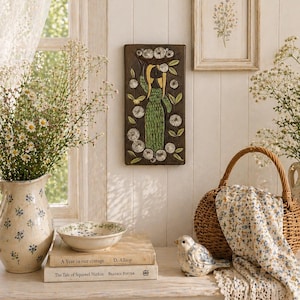 Op de afbeelding: Een serene interieurscène met een decoratieve wandplaat met een groene figuur, een bloemstuk in een vaas en een kleine vogelbeeldje. Een rieten mand en boeken dragen bij aan de cottagecore-esthetiek.