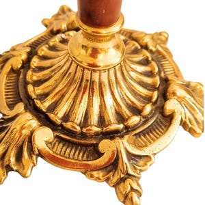 May include: Close-up of a gold-colored, ornate candlestick holder. The base features intricate scrollwork and a fluted design. The stem is a dark brown color.