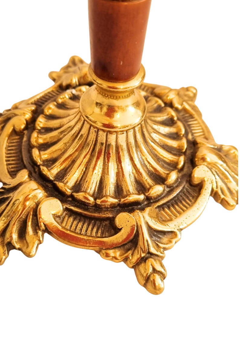 May include: Close-up of a gold-colored, ornate candlestick holder. The base features intricate scrollwork and a fluted design. The stem is a dark brown color.