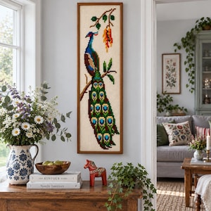 May include: A vertical rectangular needlepoint wall hanging featuring a colorful peacock perched on a branch with green leaves and orange flowers, framed in a simple brown wooden frame. The peacock has blue, green, and gold plumage.