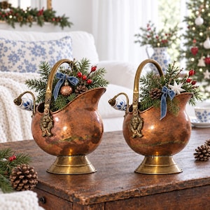 May include: Two copper-toned metal coal scuttles with brass-coloured bases and handles. Each features a lion head detail, a blue and white ceramic side handle, and is filled with evergreen branches, pinecones, red berries, and blue velvet bows.