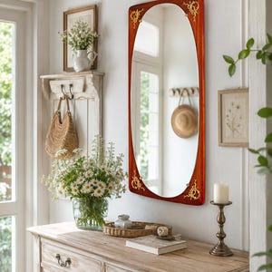 May include: A rectangular wall mirror with a polished reddish-brown wooden frame featuring rounded corners and ornate gold-toned metal accents at each corner. The mirror has a smooth, reflective surface that displays a clear view of the room interior.