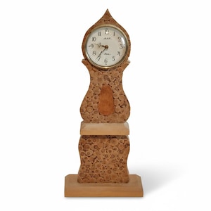 May include: A wooden grandfather clock with a light brown finish. The clock face is round with black numerals and hands. The clock's body is decorated with small, circular wooden pieces, and it stands on a rectangular base.