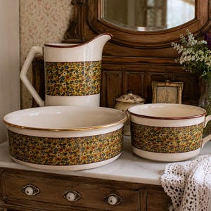 May include: A vintage ceramic pitcher, bowl, and mug set. The white pieces feature a band of floral pattern in shades of green, yellow, and red. The bowl has a gold rim. The set is displayed on a marble-topped cabinet.