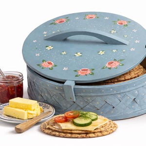 May include: A light blue, round bread basket with a floral design on the lid. The basket contains round, flatbreads. A small plate with butter, a jar of jam, and a cracker with toppings are also present.