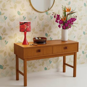 Può includere: Una consolle in legno con due cassetti, una lampada rossa con paralume fantasia, una ciotola decorativa e un vaso di fiori. La lampada è a sinistra, i fiori a destra. Il tavolo misura circa 120 cm di lunghezza.