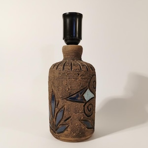 Può includere: Una lampada in ceramica marrone con portalampada nero. La lampada ha una superficie strutturata con disegni geometrici e a foglie blu. La lampada è alta circa 30 cm.