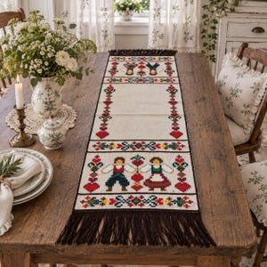 Könnte beinhalten: Ein rechteckiger, cremefarbener Tischläufer mit einem Kreuzstichmuster aus zwei Figuren in traditioneller Kleidung, roten Herzen und Blumenmotiven. Der Läufer ist an beiden Enden mit dunkelbraunen Fransen und einer Bordüre versehen.