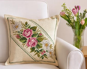 Puede incluir: Cojín decorativo color crema con un diseño floral de punto de cruz. El cojín presenta rosas rosas, flores blancas y hojas verdes dentro de un borde verde cosido. Un plato floral cuelga de la pared detrás del cojín.