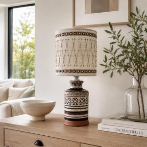 May include: A table lamp with a cylindrical ceramic base featuring horizontal bands of dark brown, white, and black geometric patterns. The drum-shaped lampshade is made of off-white fabric with gold-colored stitched diamond patterns.