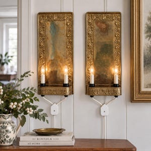 May include: A pair of rectangular wall sconces featuring an ornate gold-toned metal frame with embossed floral detailing and a weathered mirror backplate. Each sconce holds two vertical light bulbs and includes a white cord with a wall plug.