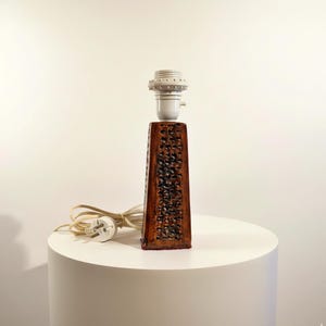 Puede incluir: Una lámpara de mesa rectangular marrón con un diseño tallado y texturizado. La lámpara tiene un casquillo blanco y un cable de alimentación con enchufe. La lámpara está sobre una superficie blanca.