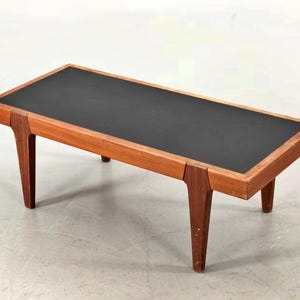 Pode incluir: Mesa de centro retangular com tampo preto e estrutura de madeira. A mesa tem quatro pernas anguladas e um acabamento escuro. A mesa tem aproximadamente 122 cm de comprimento, 51 cm de largura e 41 cm de altura.