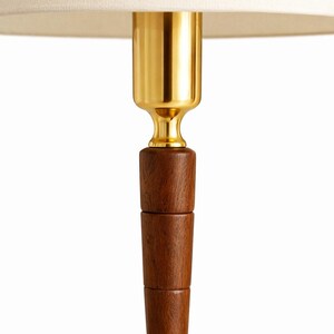May include: A close-up of a lamp base with a cream-colored lampshade. The lamp features a gold-colored metal neck and a dark brown wooden base. The wooden base is composed of stacked, cylindrical sections.