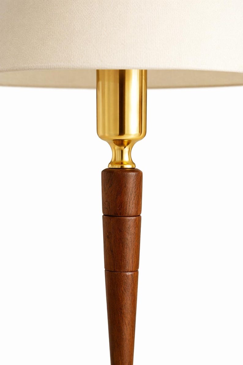 May include: A close-up of a lamp base with a cream-colored lampshade. The lamp features a gold-colored metal neck and a dark brown wooden base. The wooden base is composed of stacked, cylindrical sections.