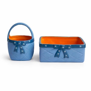 May include: Two blue ceramic baskets. One is a handled basket, the other is rectangular. Both have a diamond pattern, blue bows with white flower accents, and orange interiors. The baskets are on a white background.