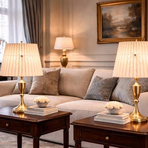 Op de afbeelding: Twee bijpassende tafellampen met geplooide kappen en gouden voetstukken staan op donkere houten bijzettafels voor een beige bank. Decoratieve kussens en een schilderij maken de elegante woonkamer compleet.
