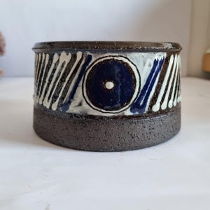 May include: A handmade ceramic bowl with a dark brown base and rim. The bowl features a central blue circle design with a white dot, and white and blue vertical stripes. The bowl has a textured, speckled appearance.
