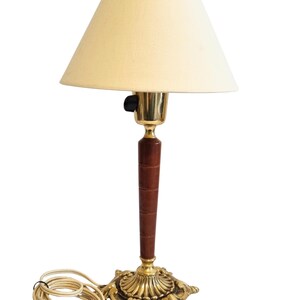 May include: A vintage table lamp with a cream-colored lampshade. The lamp features a brass base with an ornate design, a dark brown wooden stem, and a brass neck. The lamp has a power cord with a two-prong plug.