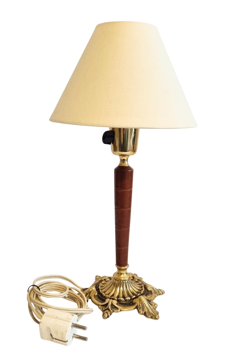 May include: A vintage table lamp with a cream-colored lampshade. The lamp features a brass base with an ornate design, a dark brown wooden stem, and a brass neck. The lamp has a power cord with a two-prong plug.