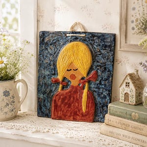 May include: A square ceramic wall plaque featuring a relief sculpture of a person with long yellow hair in pigtails, a red shirt, and closed eyes. The background is a textured, dark blue glaze. A light-colored rope loop is attached to the top for hanging.