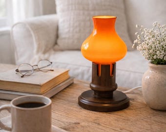 Puede incluir: Una lámpara de mesa con una pantalla de cristal de color naranja vibrante y silueta suave y redondeada. La pantalla se apoya sobre una base de madera circular de color marrón oscuro con un diseño de pilar central. Emite una luz cálida.