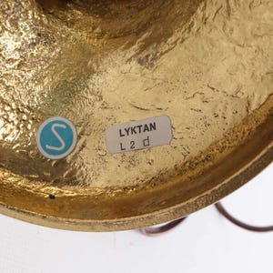 Op de afbeelding: Close-up van een messingkleurig metalen object met een getextureerd oppervlak. Een blauw-witte sticker met een "S" is zichtbaar, samen met een label met de tekst "LYKTAN L 2 d". Het object lijkt deel uit te maken van een groter decoratief stuk.
