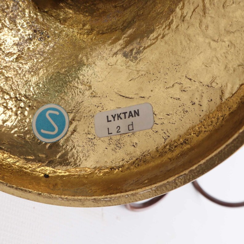 Op de afbeelding: Close-up van een messingkleurig metalen object met een getextureerd oppervlak. Een blauw-witte sticker met een "S" is zichtbaar, samen met een label met de tekst "LYKTAN L 2 d". Het object lijkt deel uit te maken van een groter decoratief stuk.