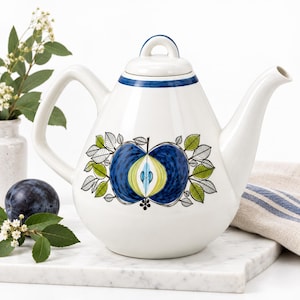 May include: A white ceramic teapot featuring a blue and green apple illustration on the front. The teapot has a curved handle, a matching lid with a loop handle, and a blue band around the neck. The apple design is surrounded by green leaves.