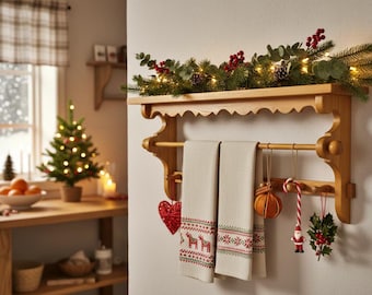 Puede incluir: Estante de madera con dos toallas beige con bordados rojos y verdes. El estante está adornado con vegetación, luces y adornos navideños. Un pequeño árbol de Navidad y naranjas son visibles en el fondo.
