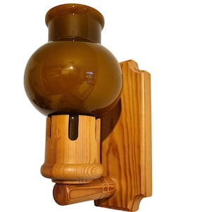 May include: A wall-mounted light fixture featuring a rounded, amber-colored glass shade secured within a light-toned wooden base. The wooden mount has a vertical rectangular backplate with a cylindrical arm extending forward to support the glass globe.