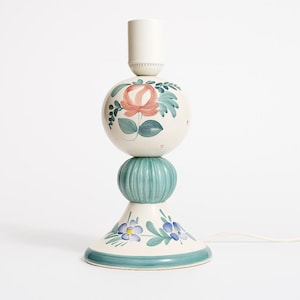 May include: A ceramic table lamp with a floral design. The lamp features a cream-coloured base with blue and teal accents, a teal-coloured centre, and a cream-coloured top with a pink rose and green leaves. The lamp is approximately 36 cm tall.